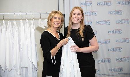 A nursing instructor hands a white coat to a student.